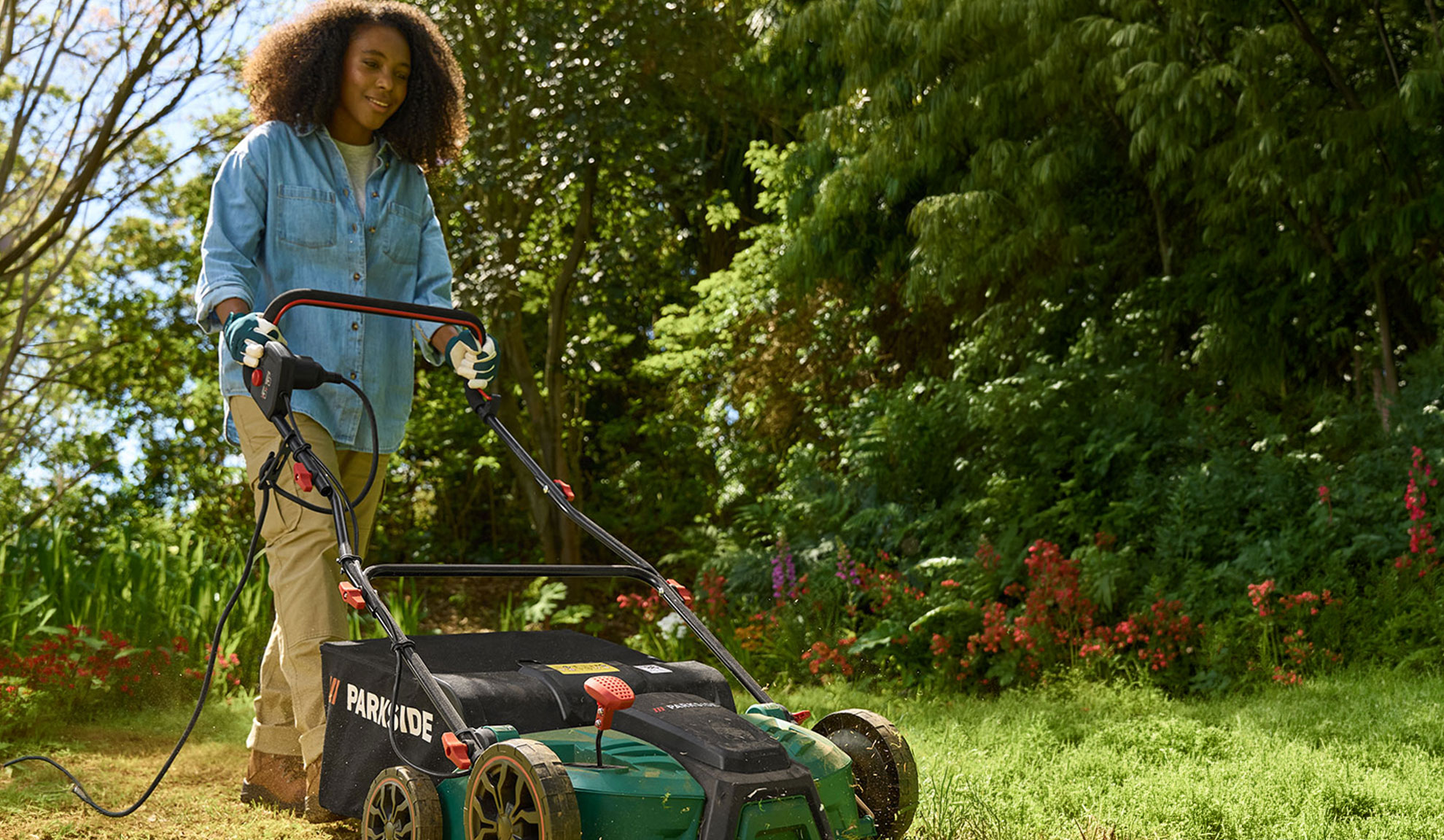 A woman in a denim shirt and khaki pants mows a lawn with an electric lawnmower.