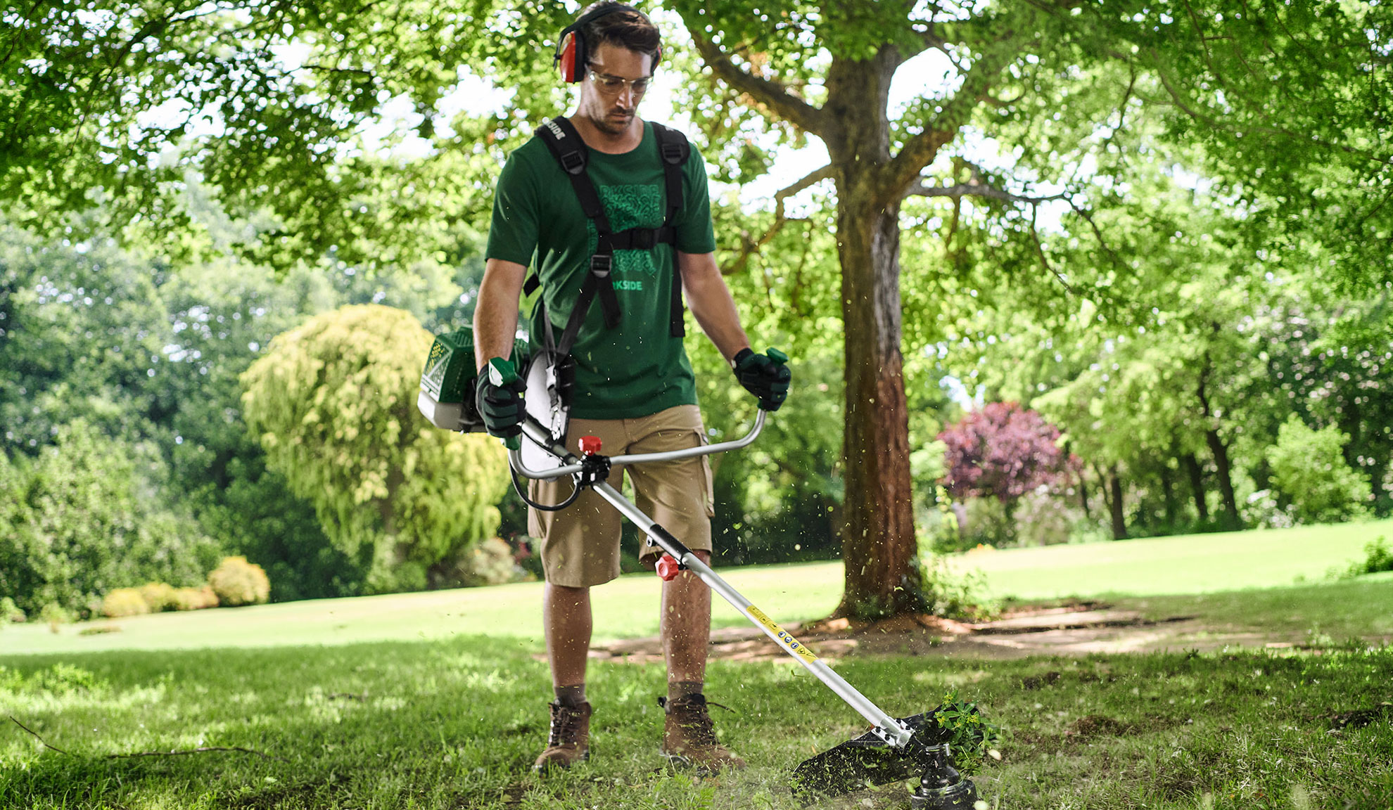 Man trimming grass in a garden with a brush cutter, wearing safety glasses and ear defenders.