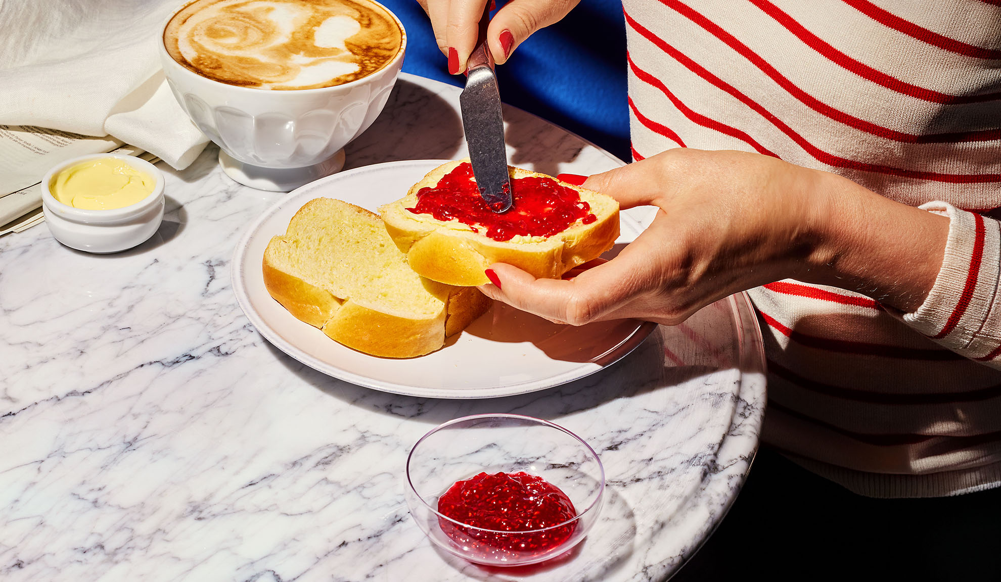 Hands spreading jam on brioche bread, with coffee and butter on a marble table.