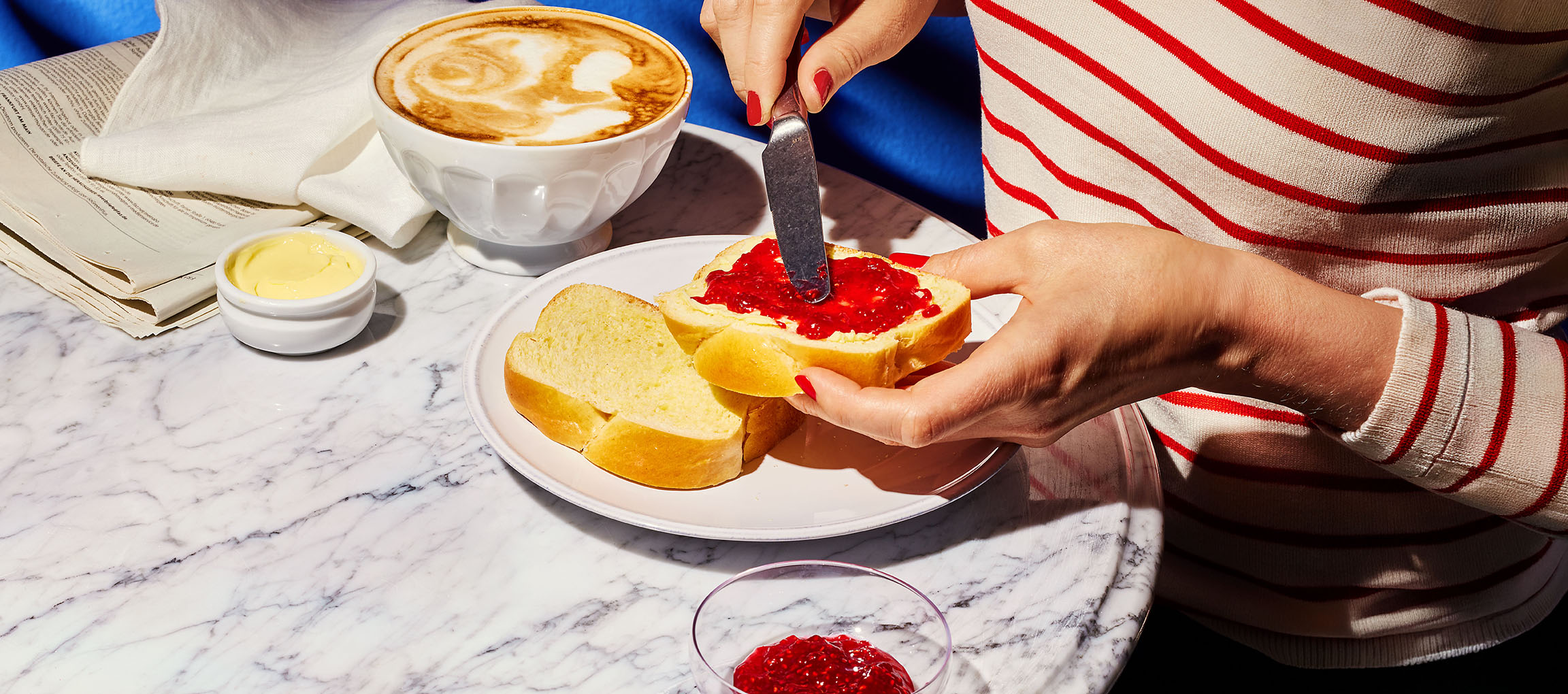 Hands spreading jam on brioche bread, with coffee and butter on a marble table.