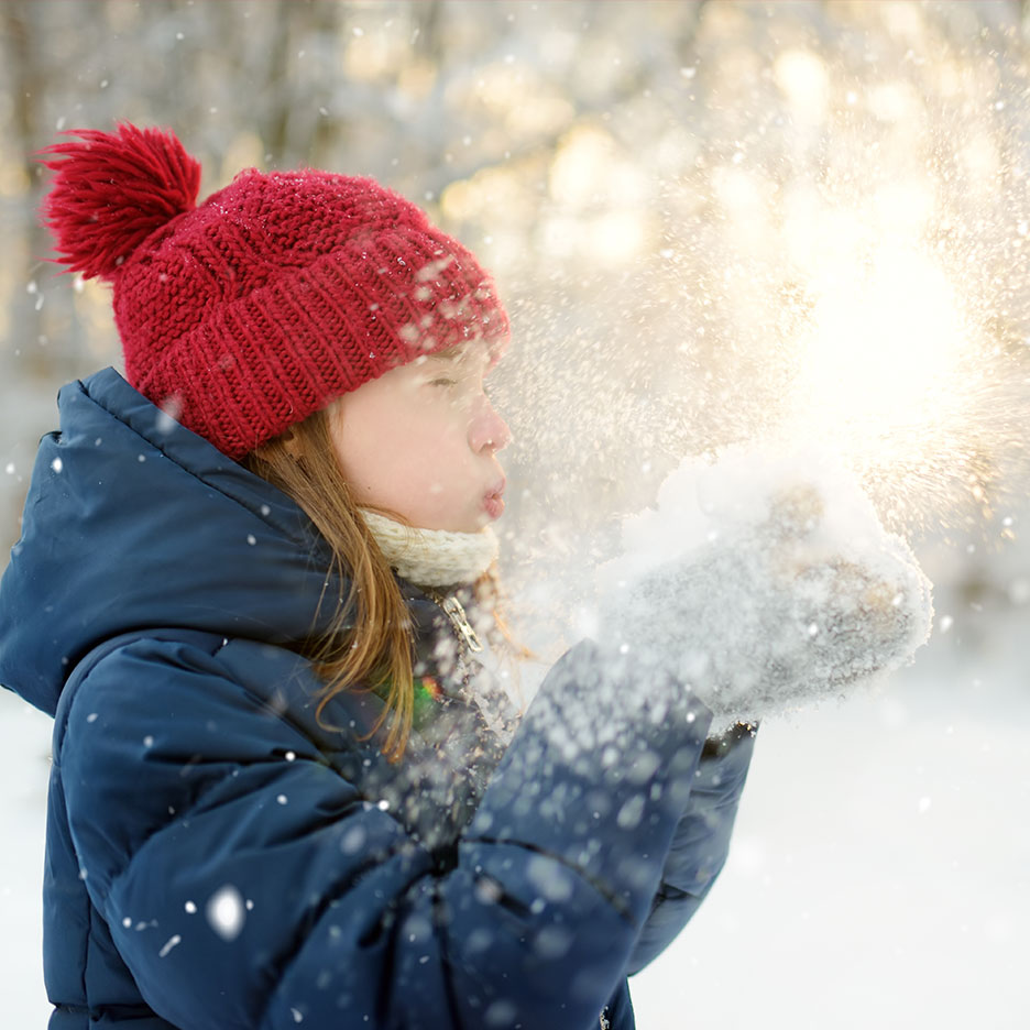 Child in red hat and blue jacket blowing snow in sunshine.
