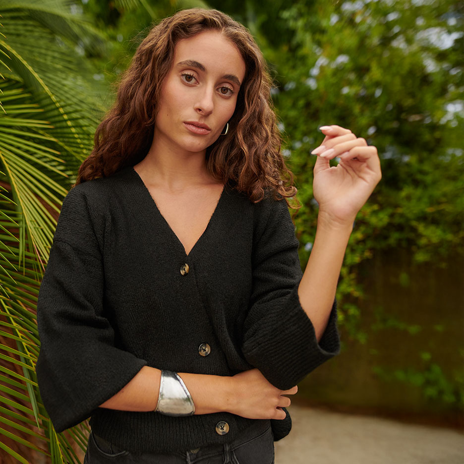 Woman in black button-up cardigan and silver cuff bracelet in front of green plants.