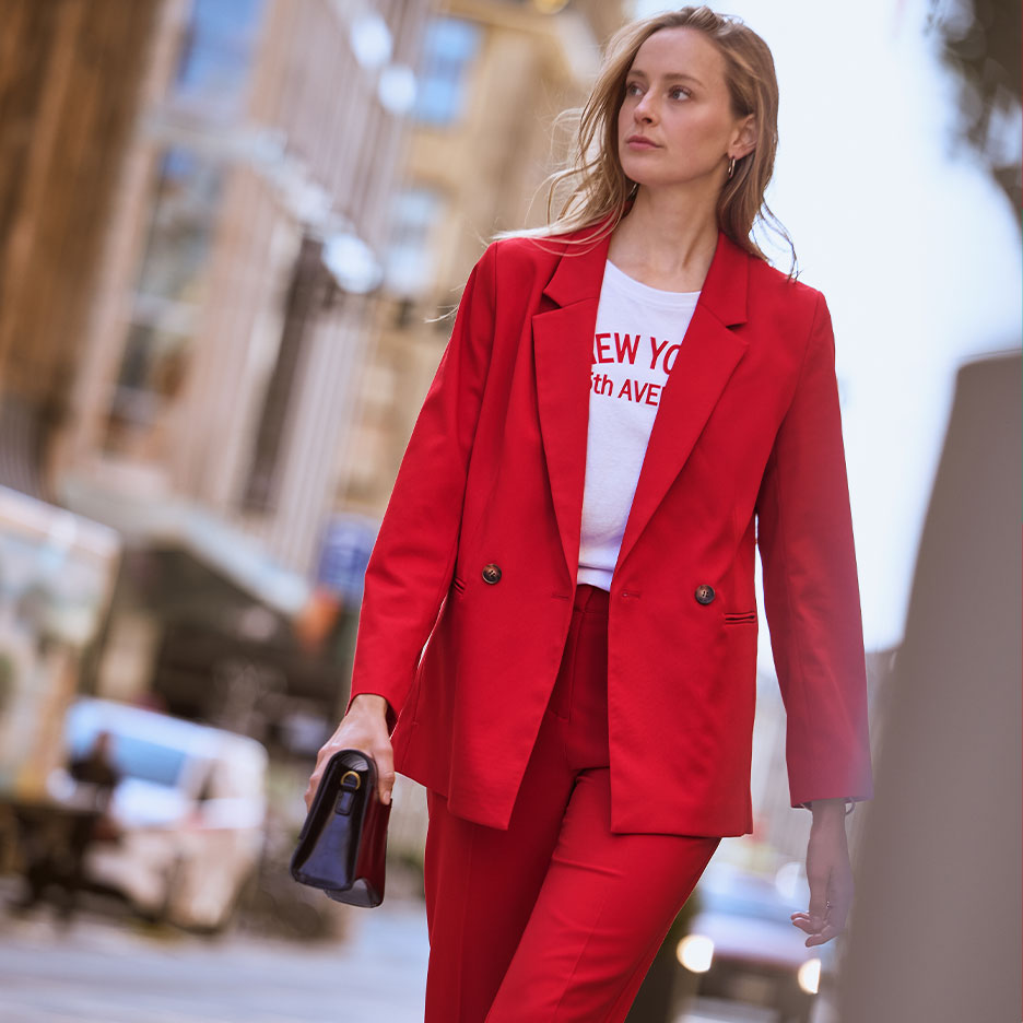 Woman in a red pantsuit and white t-shirt with 'NEW YORK 5th AVE' text, holding a black handbag.