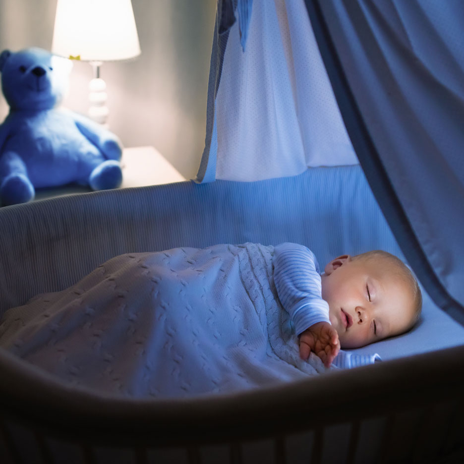 Baby sleeping peacefully in a crib with a teddy bear and lamp in the background.