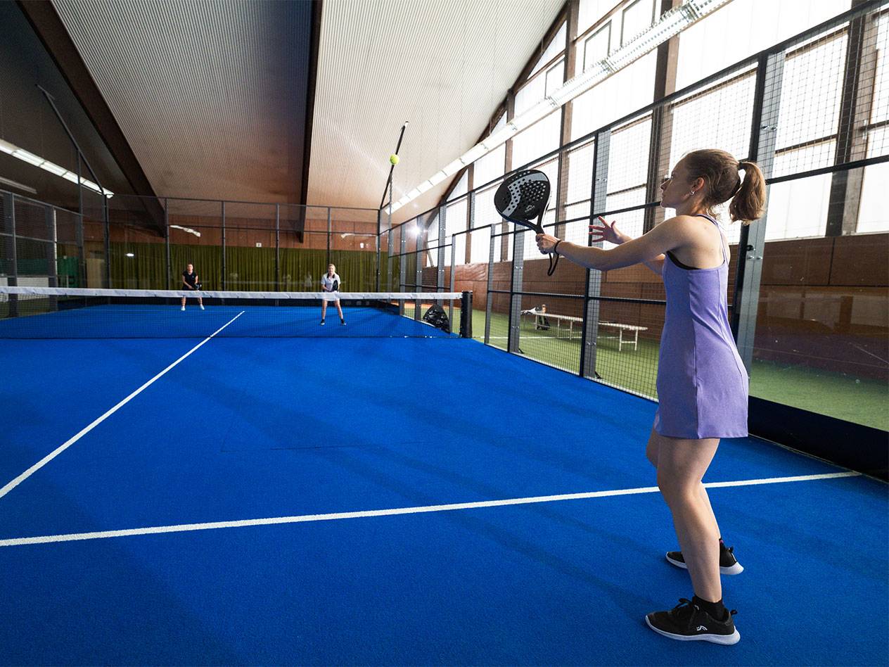 Woman in a purple dress playing padel on a blue court, other players in the background.