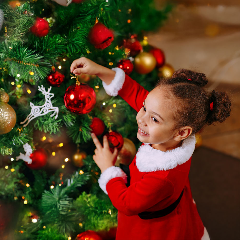 Happy girl in Santa costume decorating a Christmas tree with red baubles.