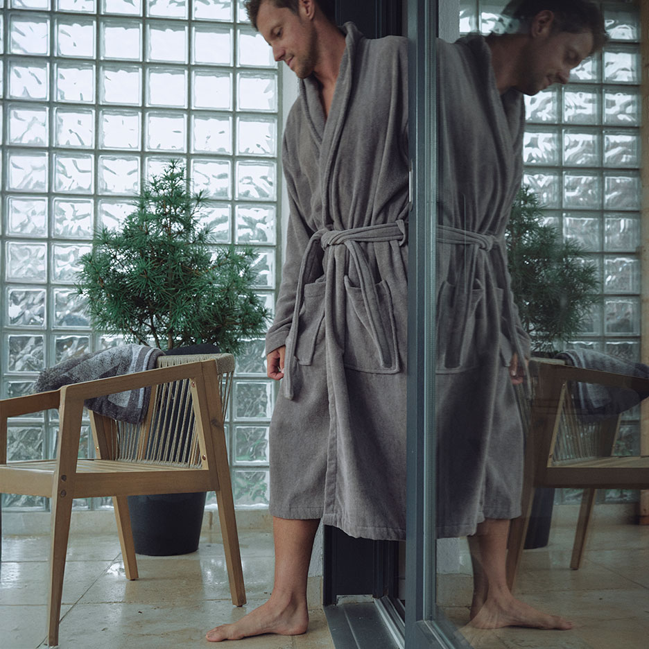 Man in grey bathrobe stands by glass block wall with a plant and chair.