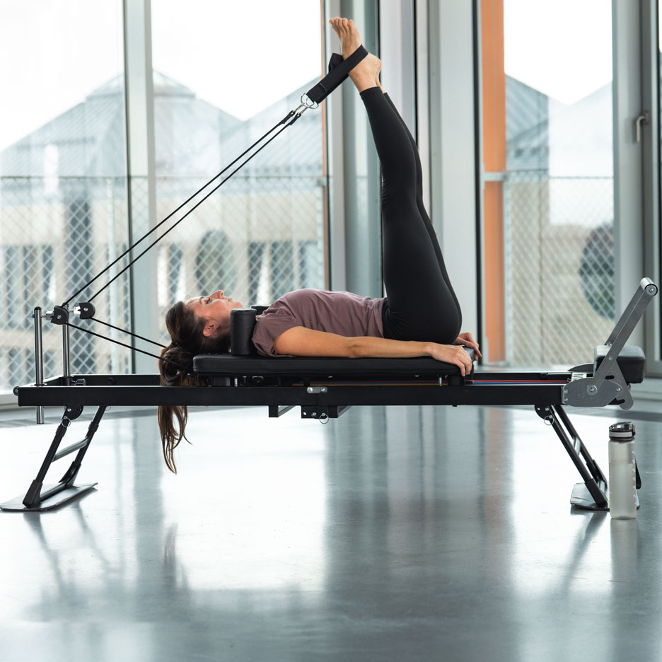 Woman doing Pilates on a reformer with legs up, in a bright room.