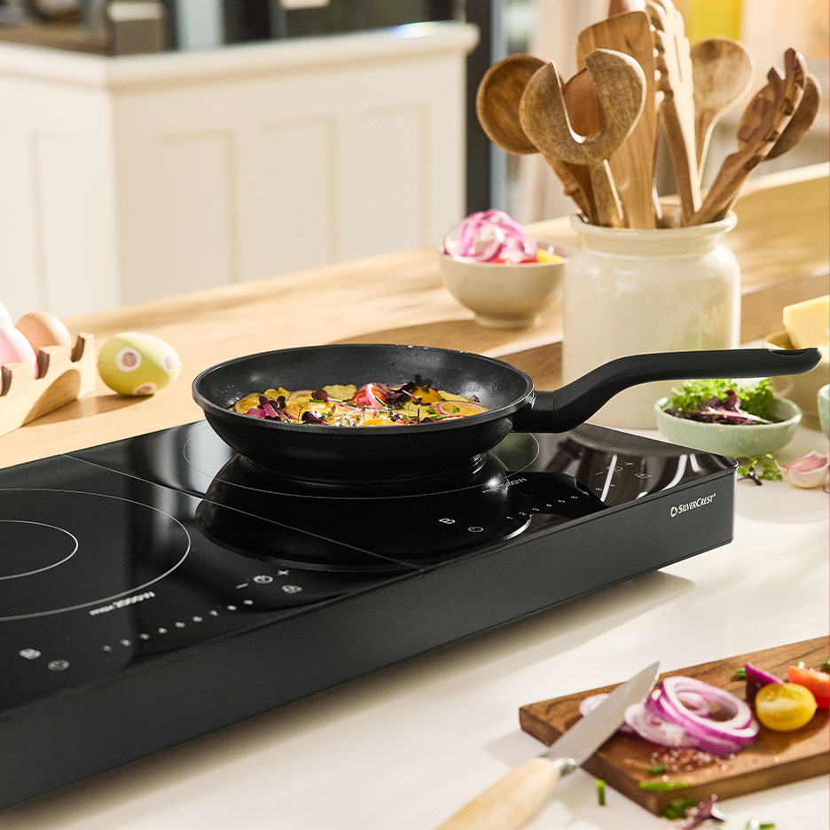 SilverCrest pan on an induction hob with vegetables and kitchen utensils.