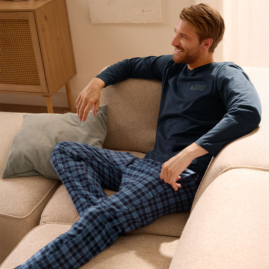 Man in dark blue long-sleeved T-shirt with 'WKND' and plaid pajama pants sitting on a sofa.