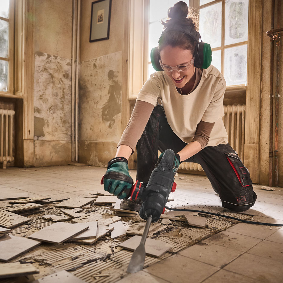 Woman with ear protection and glasses using a hammer drill to remove tiles.