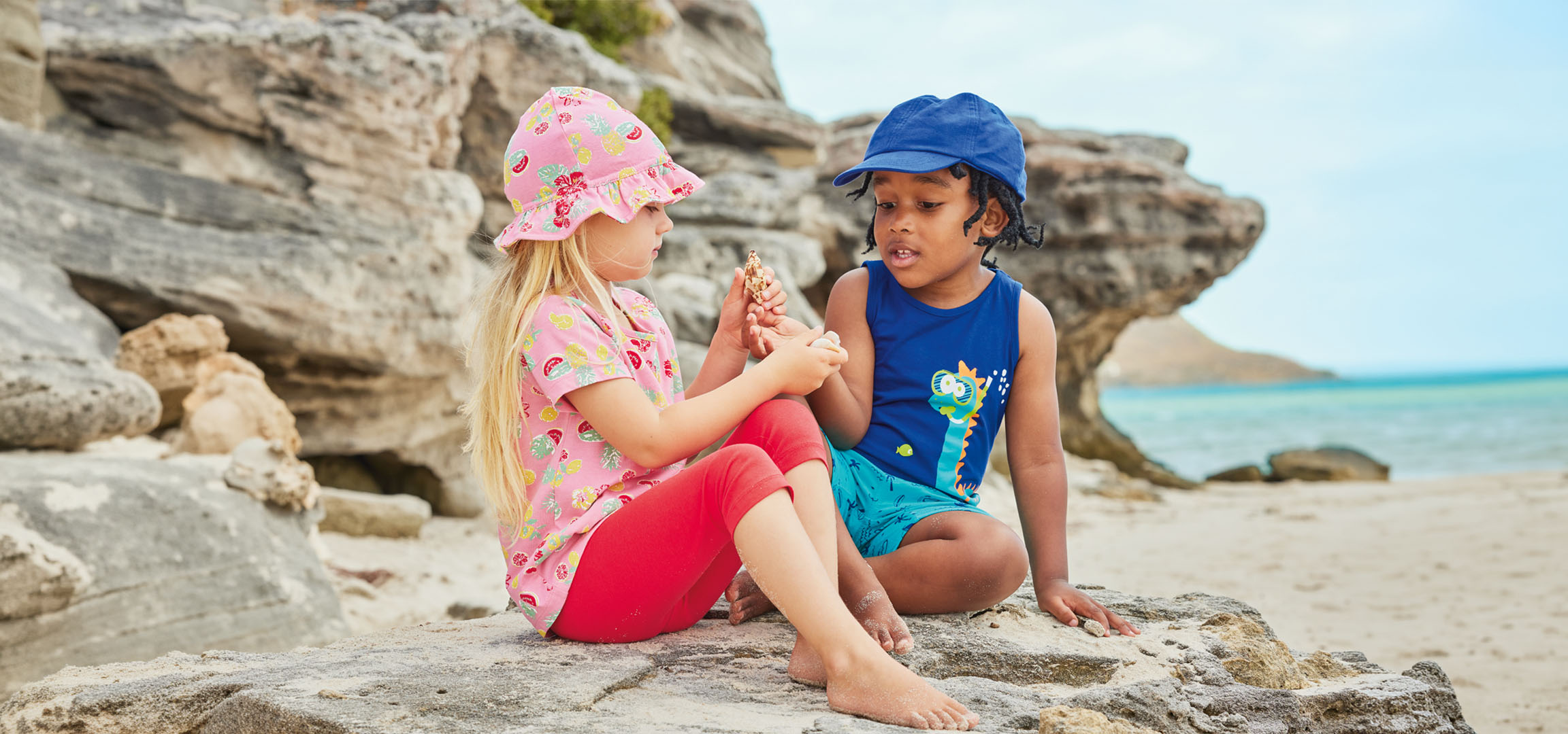 Two children in summer clothing sit on a rock by the beach, holding a seashell.