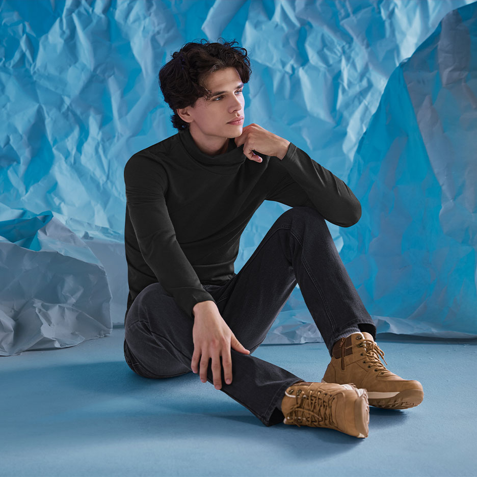 Man in black turtleneck, dark jeans, and brown boots, sitting against a crumpled blue background.