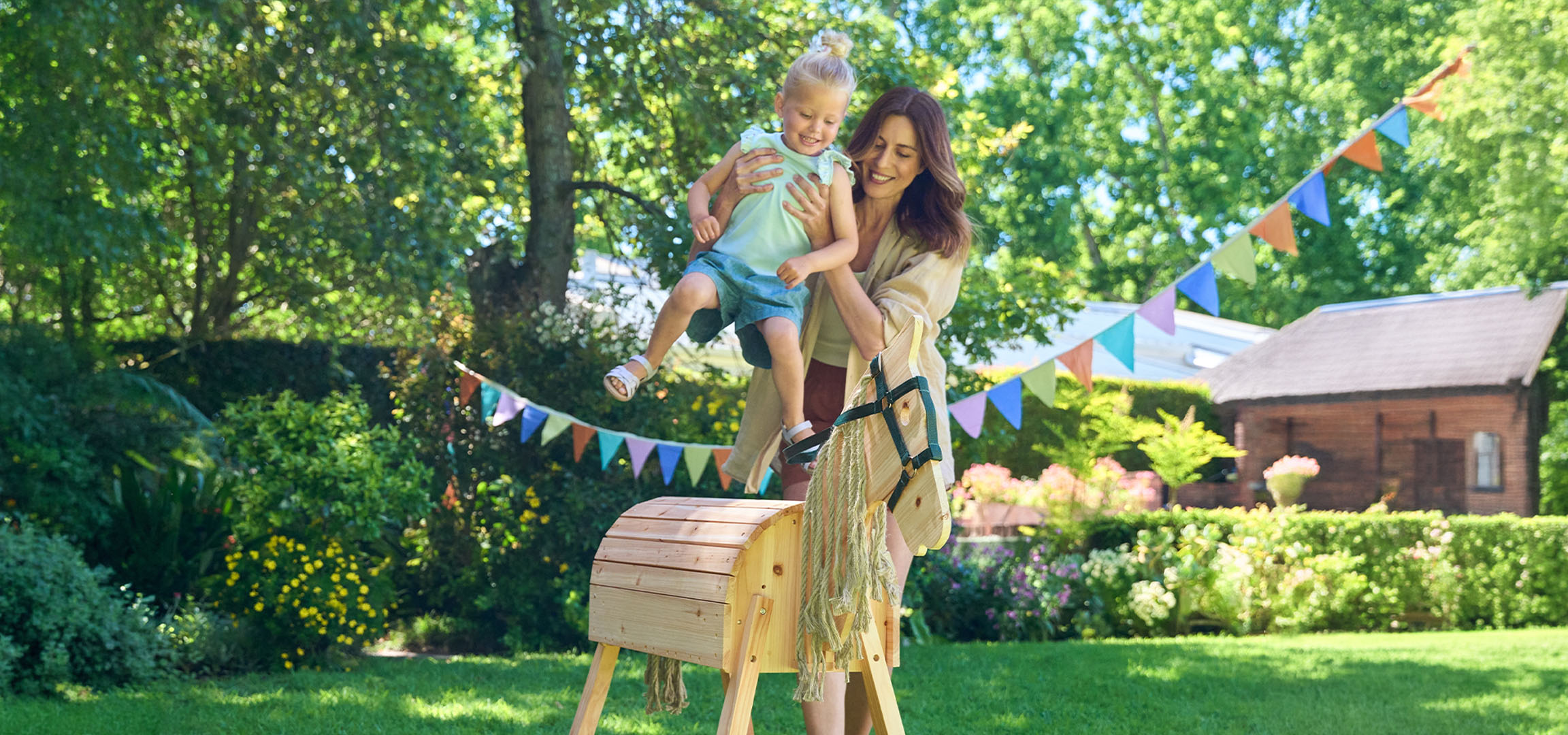 Mother and child playing with a wooden rocking horse in a sunny garden.