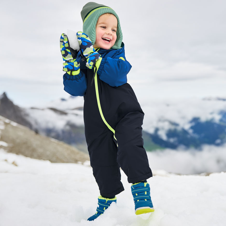 Happy child in blue snowsuit, green hat, and mittens, playing in the snow.