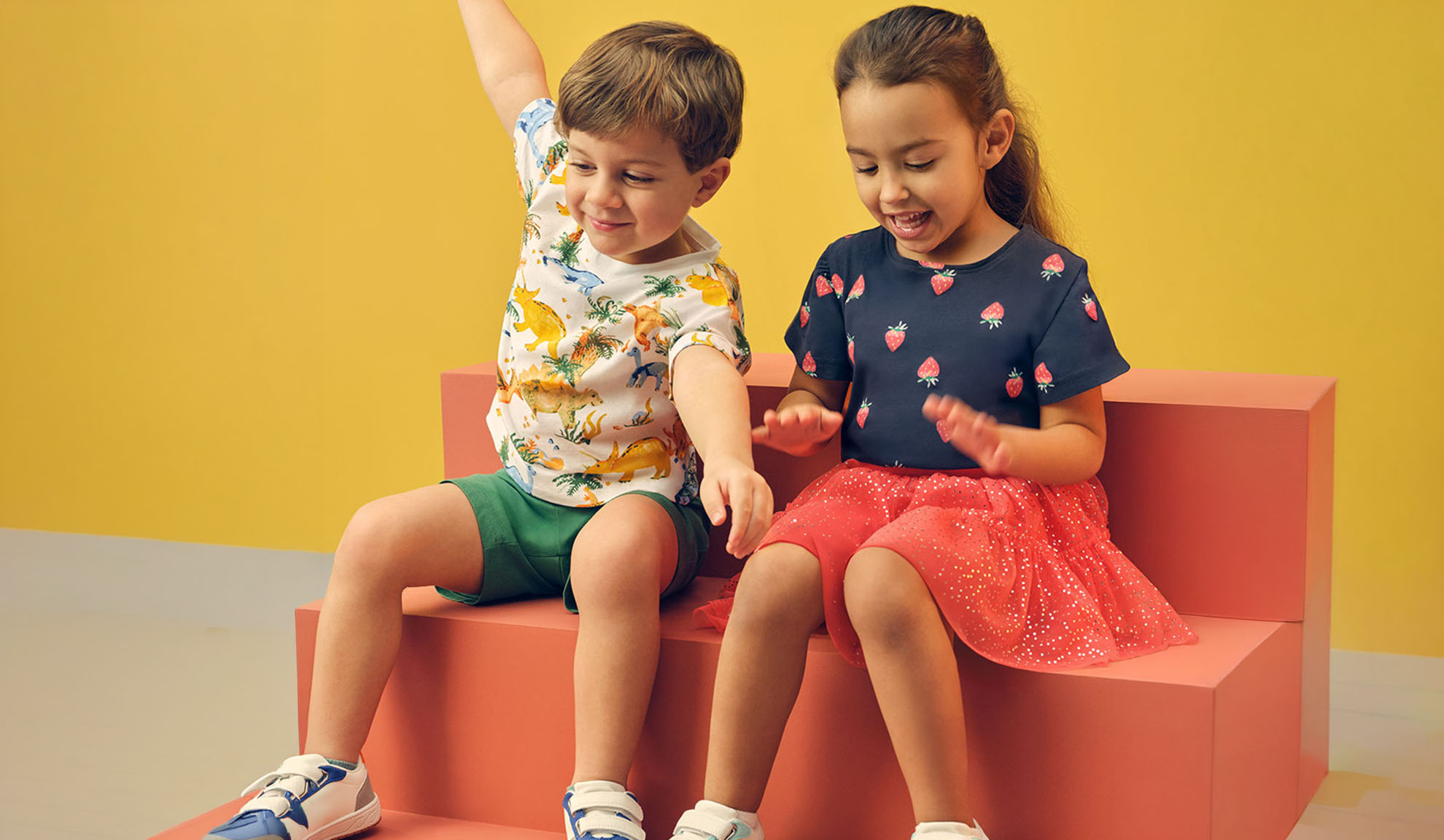 Two children in summer clothes, a boy in a dinosaur t-shirt and a girl in a strawberry t-shirt.