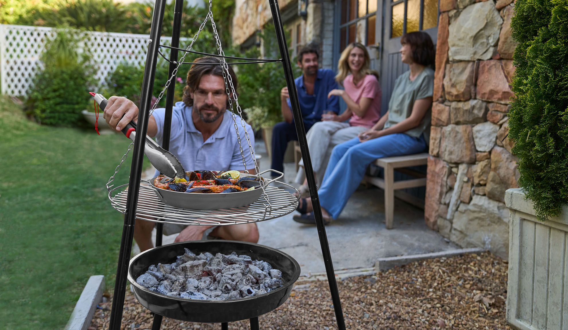 Man grilling seafood on a tripod grill, friends relaxing in the background.