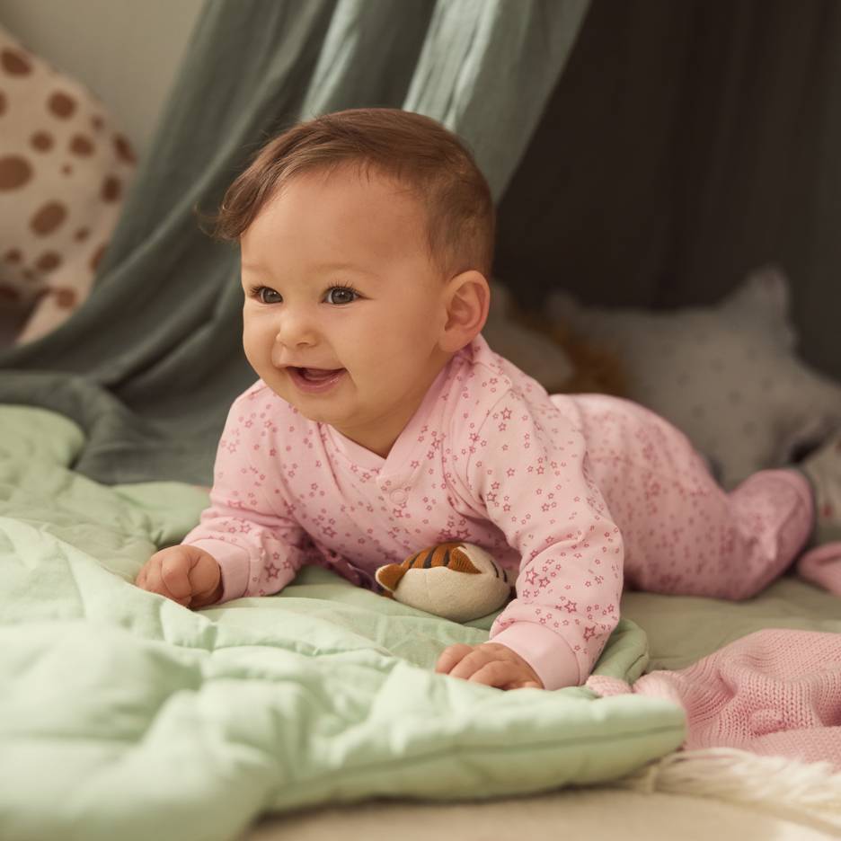 Baby in pink star pajamas, lying on a green blanket with a plush toy.